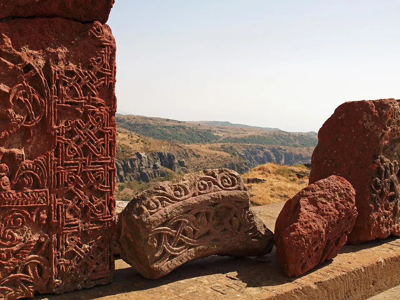 Fragments of red tuff carved with interlaced Armenian ornament and a khachkar cross-stone lying near the edge of the gorge at Amberd