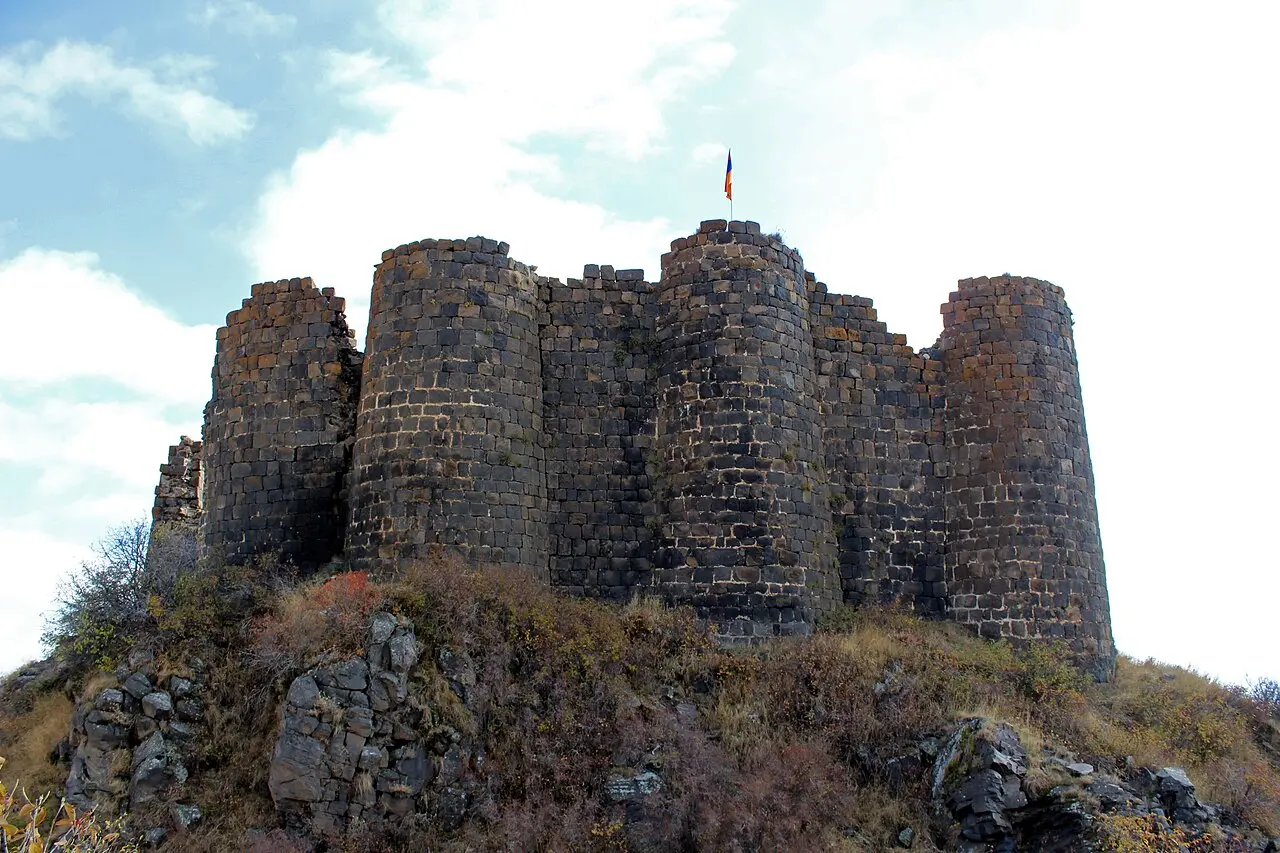 The round basalt towers and high curtain wall of Amberd fortress rising from a rocky bluff, an Armenian flag flying from the keep