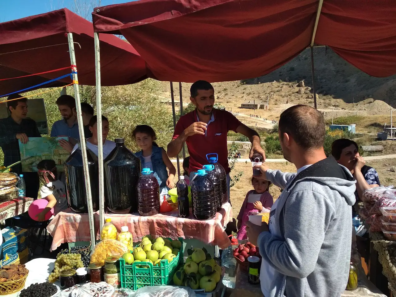 Demijohns of homemade wine and crates of fruit on a stall under a red canopy at the Areni Wine Festival