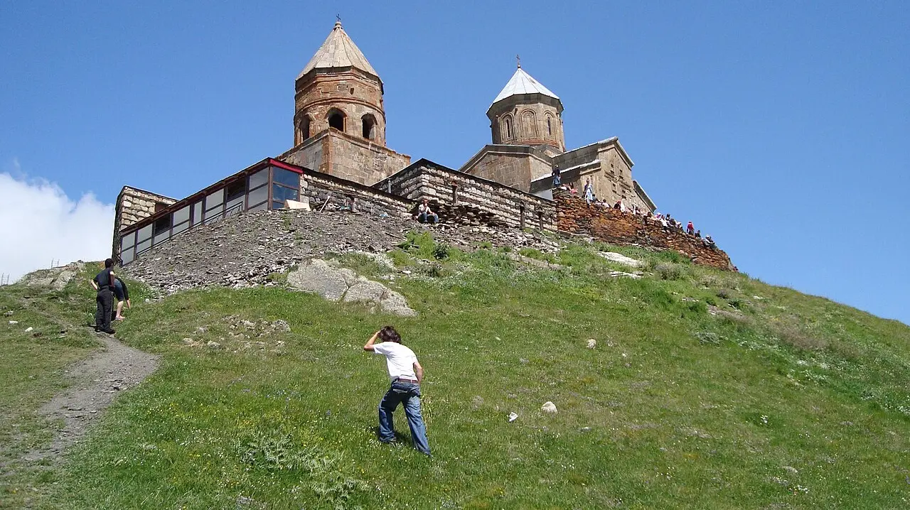 The twin stone churches of Gergeti Trinity on a green hilltop under a blue sky in the Georgian high Caucasus, with a child running up the slope