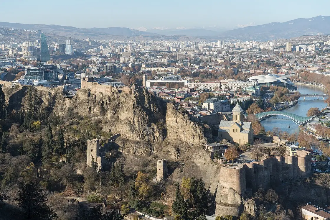 A view over Tbilisi with the Narikala fortress walls on the rocky ridge, a church, the Bridge of Peace over the Kura river and the snow-capped Caucasus behind