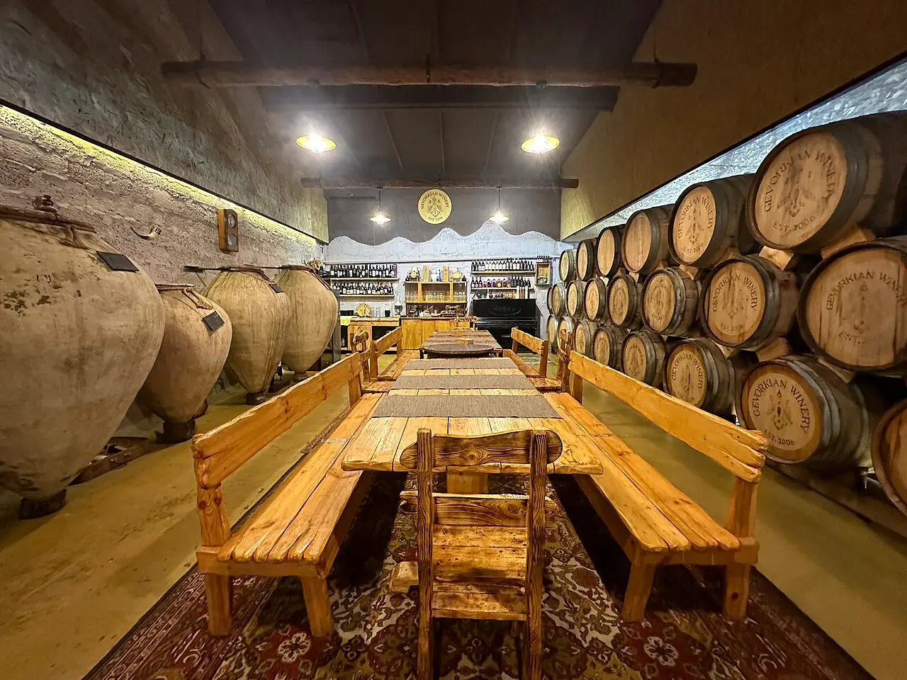 The cellar tasting room of an Armenian winery, with tall clay karas wine jars along the left wall and stacked oak barrels on the right