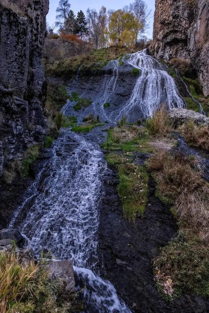 The Jermuk waterfall running down a dark cliff with autumn-yellow trees above it