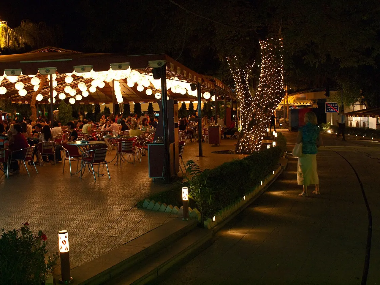 An evening outdoor cafe in central Yerevan strung with round white lanterns and a fairy-lit tree, diners at tables and a person walking past