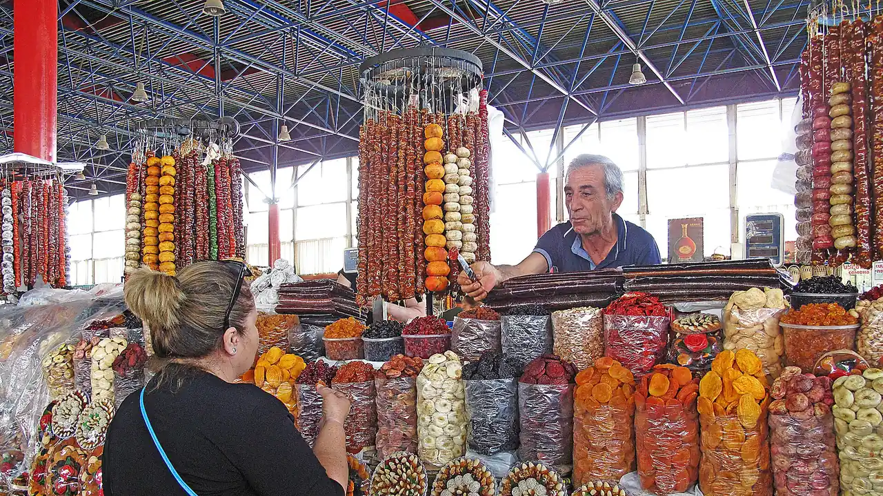 A dried-fruit vendor and a woman shopper facing each other across a colourful stall of dried apricots, nuts and churchkhela strings at a Yerevan market