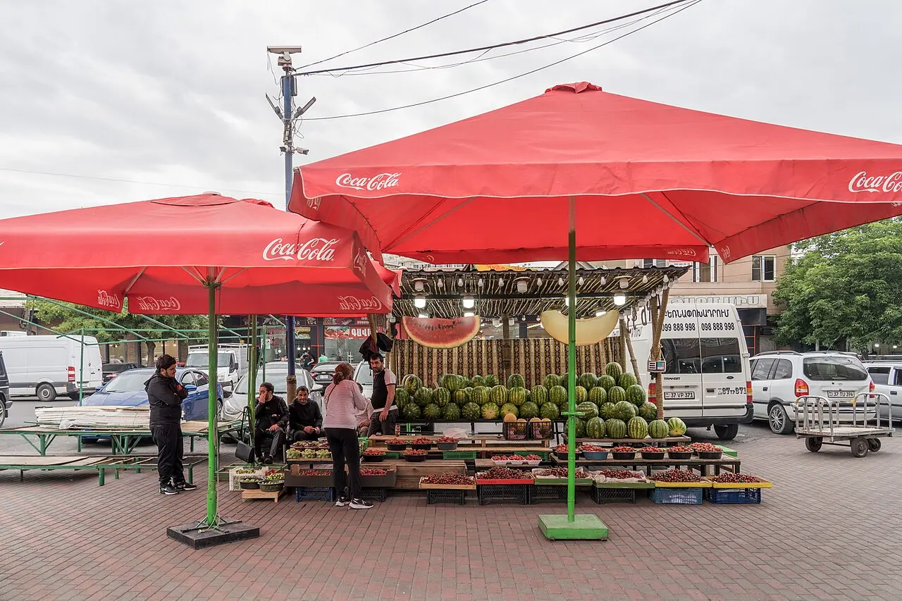 An outdoor watermelon and cherry stall under bright red umbrellas at the GUM Market in Yerevan, vendors sitting beside stacked melons