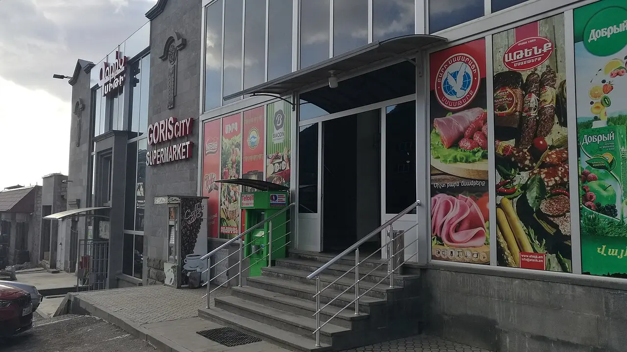 The entrance to Goris City Supermarket, a modern glass-fronted store with product posters and a payment-terminal kiosk by the steps