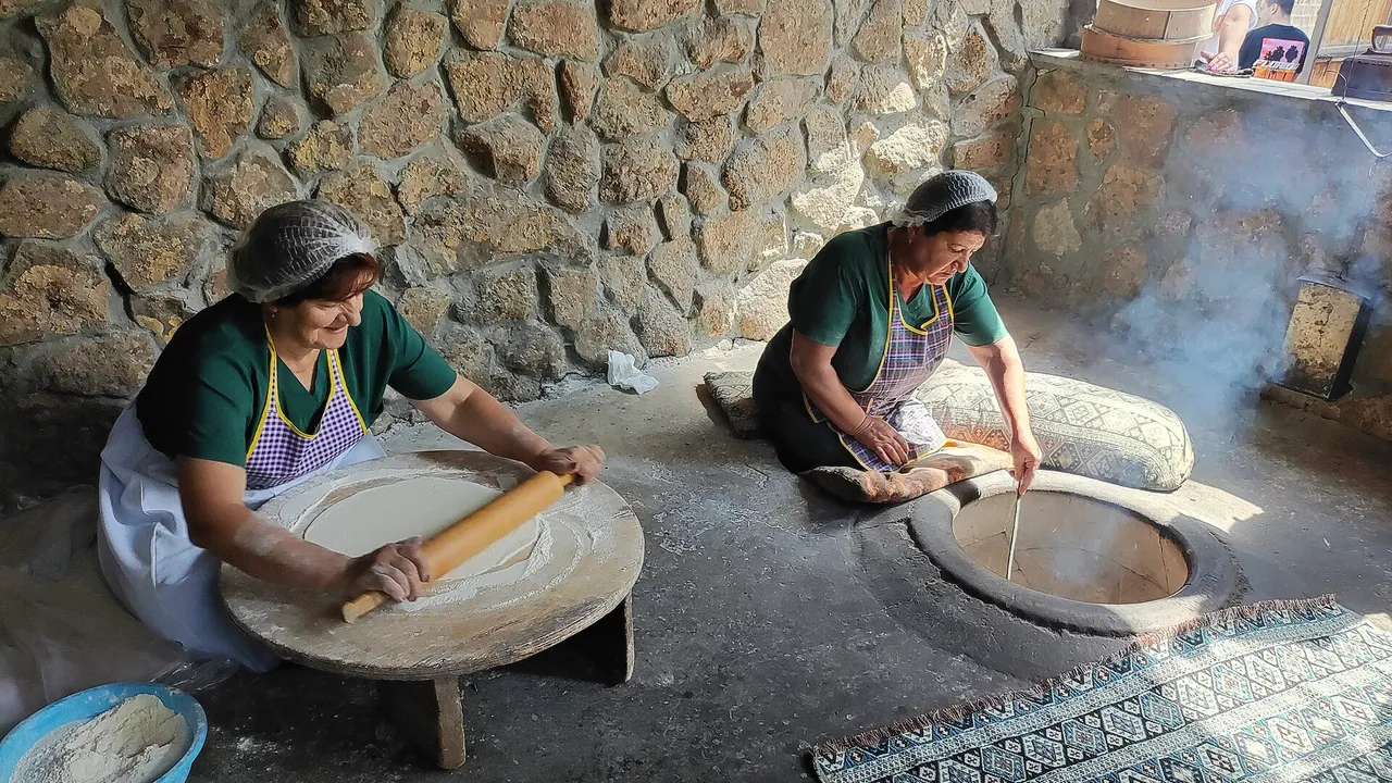 Two women baking lavash, stretching thin dough beside a sunken clay tonir oven