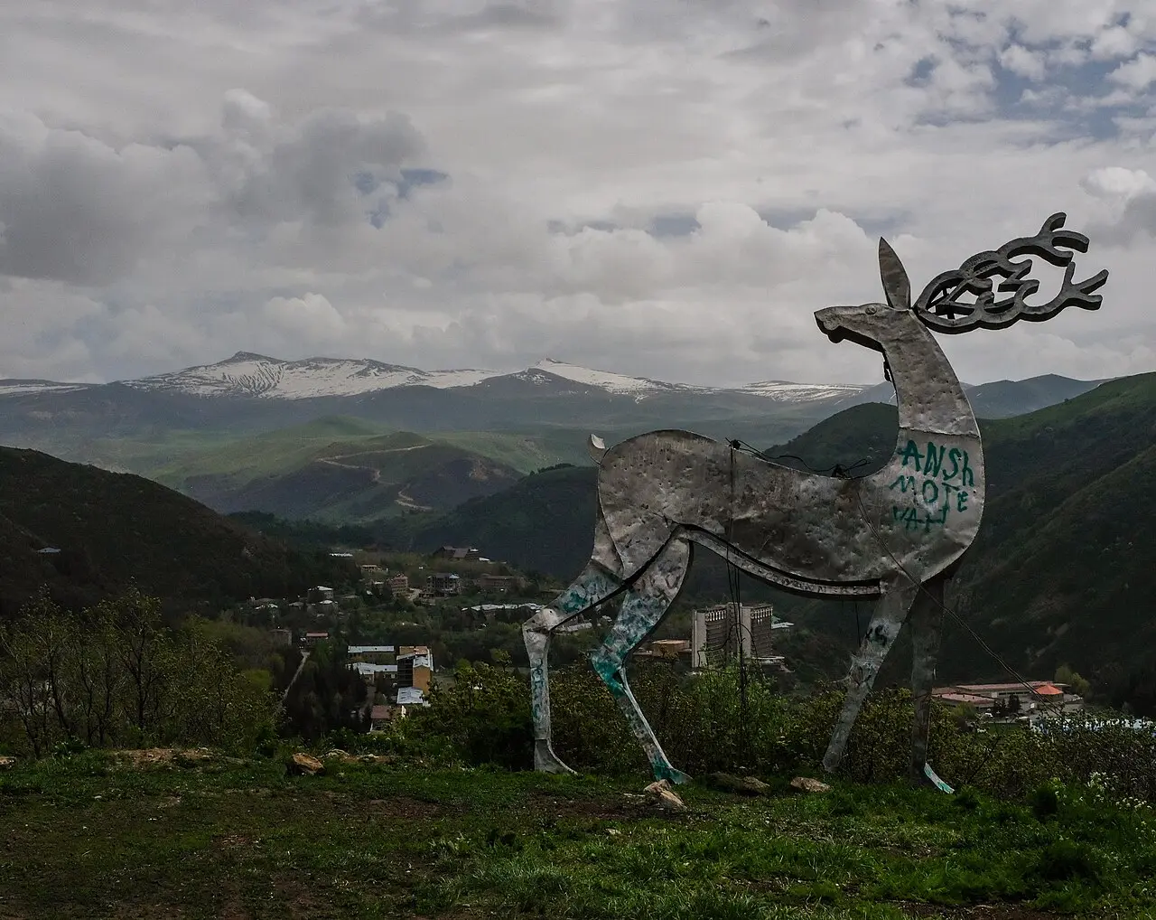 A metal deer statue on a hill overlooking the spa town of Jermuk with snow-capped mountains behind