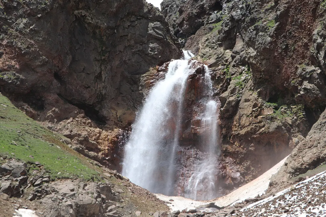 The Gegharot waterfall pouring over dark red volcanic rock on Mount Aragats with patches of old snow still lying at its base