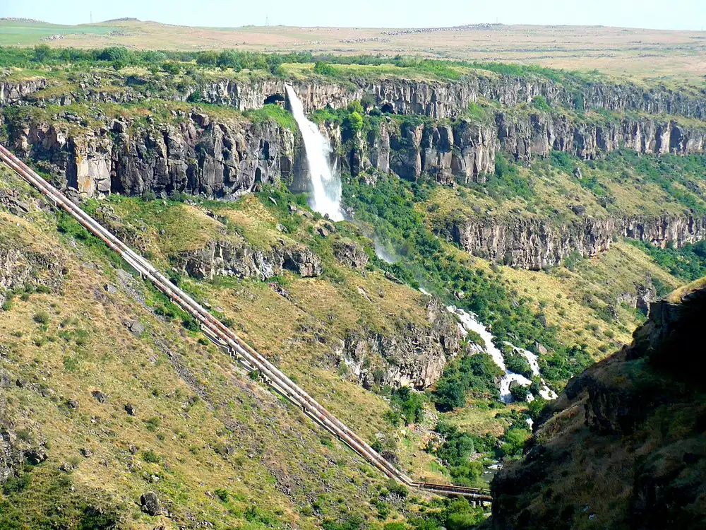 The Kasakh waterfall falling from a basalt rim into a green gorge, with two large steel pipelines running down the near slope