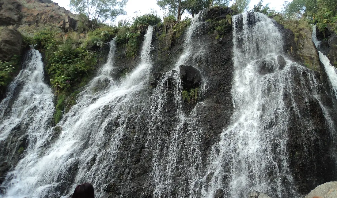 The Shaki waterfall at full flow, dozens of separate white threads fanning across a wide dark basalt ledge topped with greenery