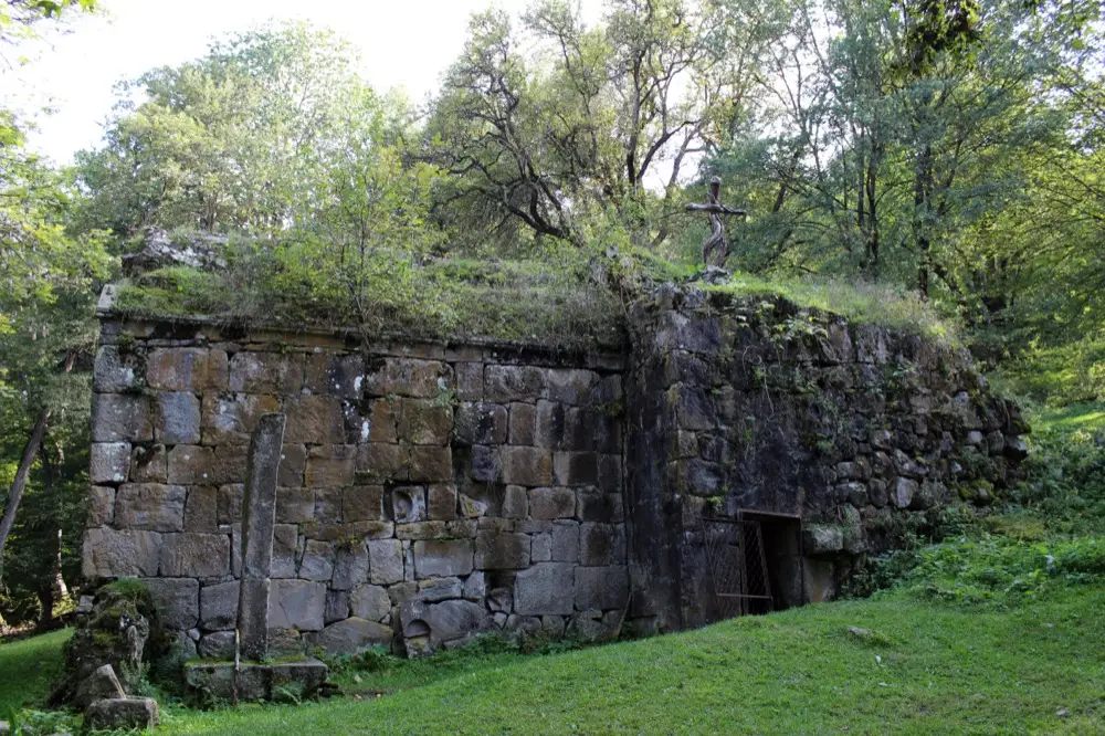 The overgrown stone ruin of Matosavank monastery, forest reclaiming its walls with a metal cross on the roofline