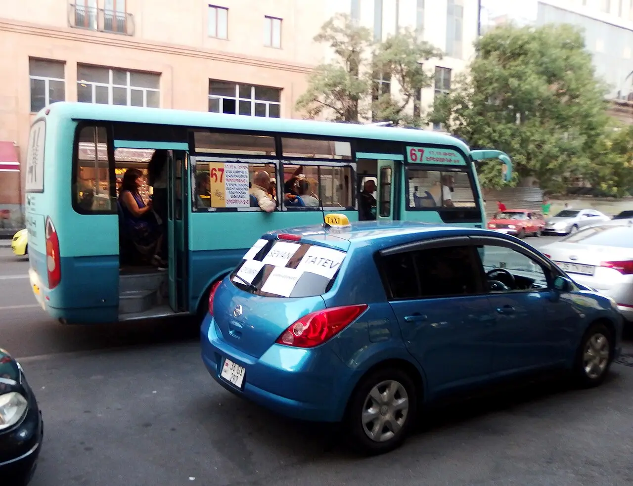A turquoise marshrutka minibus and a blue taxi with a handwritten TATEV sign parked in traffic in Yerevan