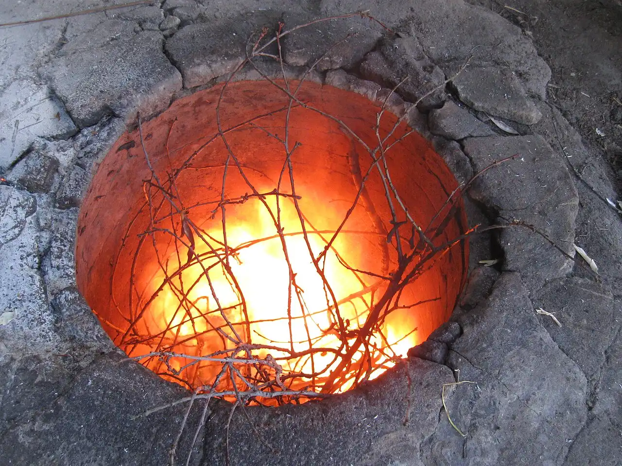 A traditional Armenian tonir clay oven sunk into the ground, glowing with fire and vine cuttings, ready for baking lavash
