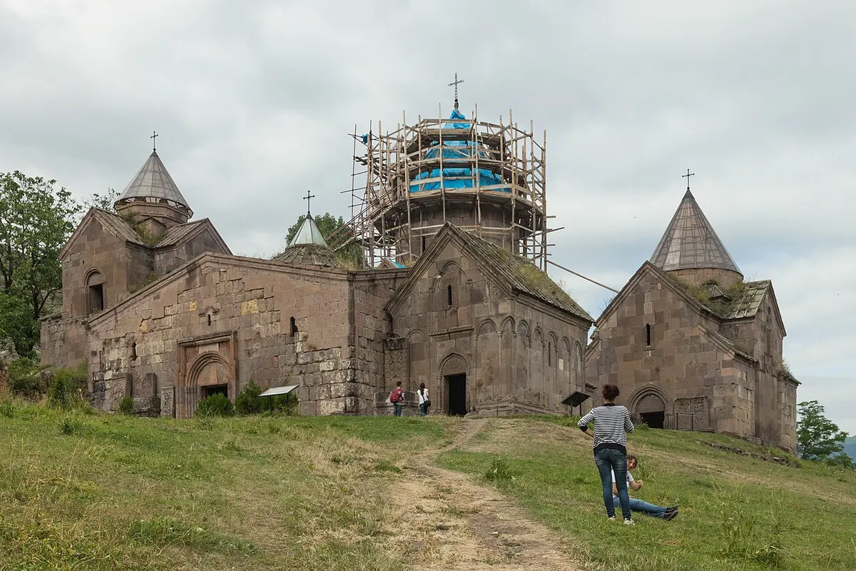 Visitors walking up a grassy hillside path toward the stone churches of Goshavank monastery, one dome wrapped in scaffolding and blue sheeting
