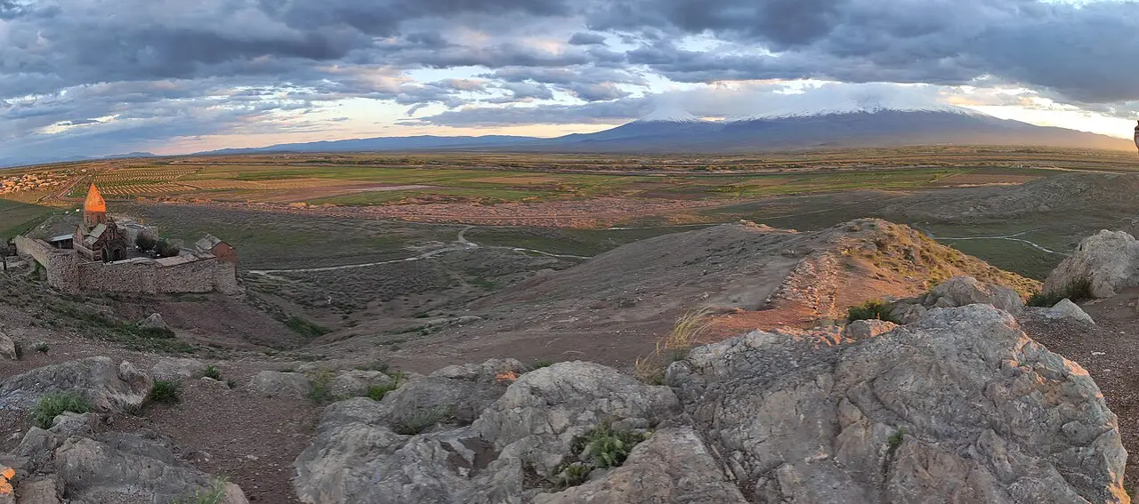 A wide dusk panorama of Khor Virap monastery on its low hill with the vast Ararat plain and the snow-capped cone of Mount Ararat behind it