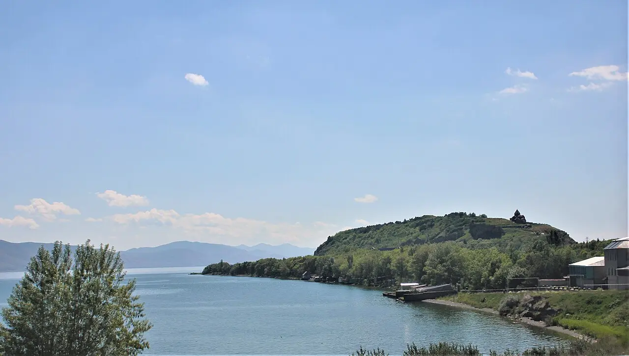 A calm view across the blue water of Lake Sevan to the green Sevan peninsula, with the small Sevanavank monastery on the hilltop in the distance