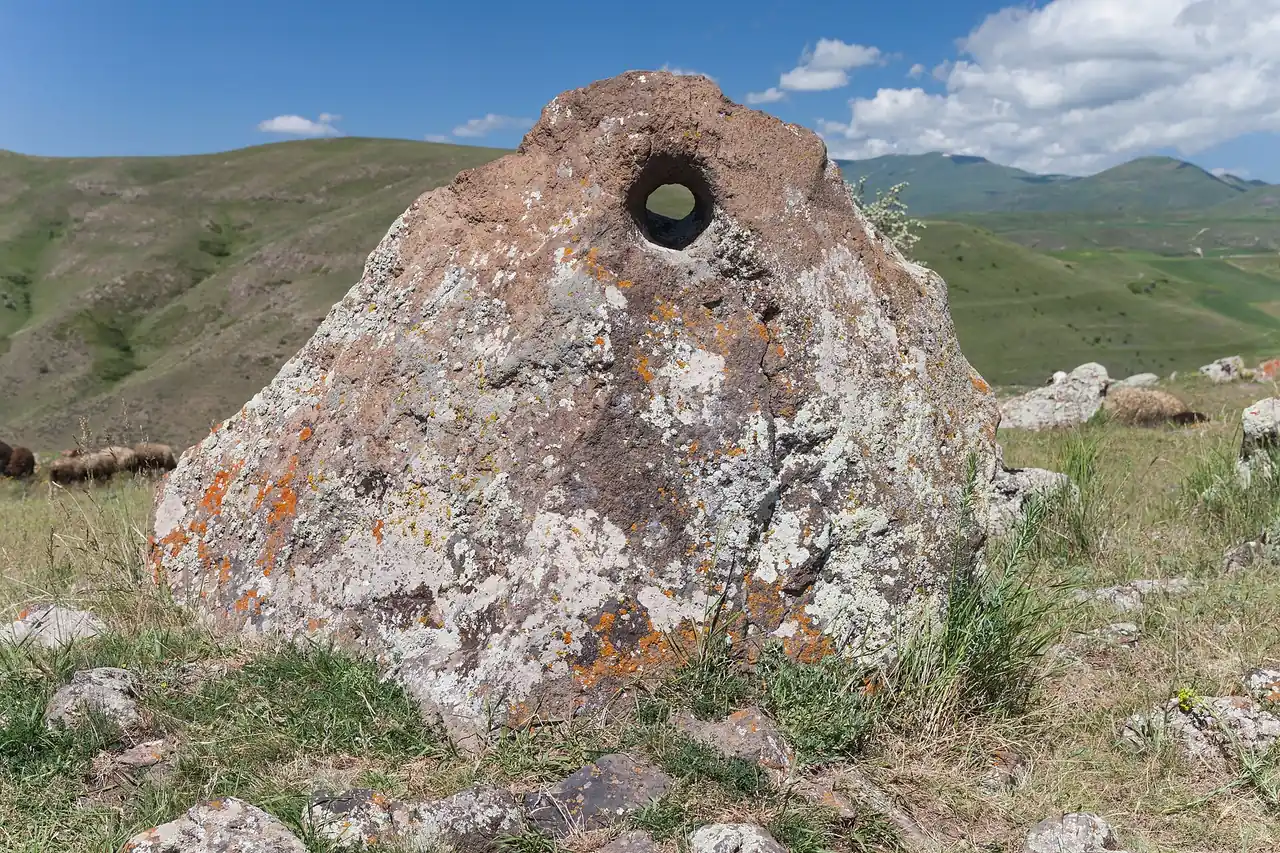 A lichen-covered standing stone with a bored round hole near its top at Karahunj, the green Syunik plateau and Zangezur mountains behind