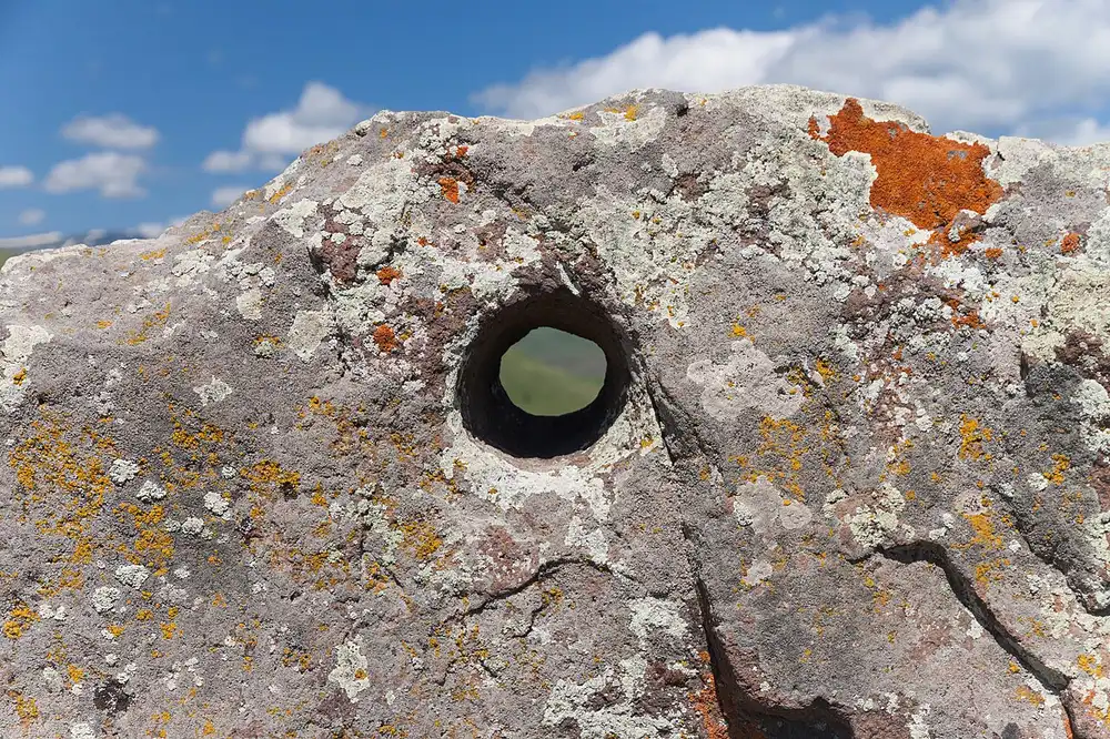 Close-up of a smooth round hole bored through a lichen-covered stone at Karahunj, framing the green valley behind