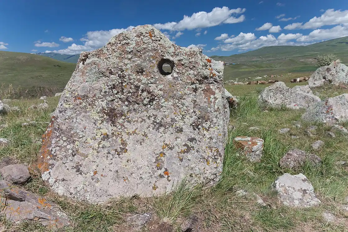 A tall pitted monolith at Karahunj with a hole near its crown, snow-streaked mountains and grazing sheep in the distance