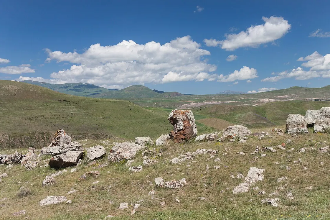 A row of upright prehistoric stones spread across the open Karahunj plateau with rolling green hills and mountains beyond