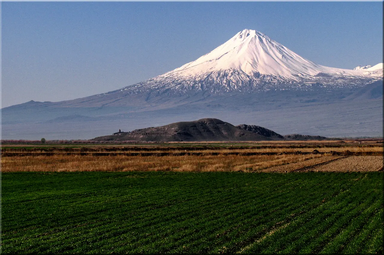 Greater Ararat under fresh snow rising above the low hill of Khor Virap monastery, seen across green and ploughed fields on a clear day