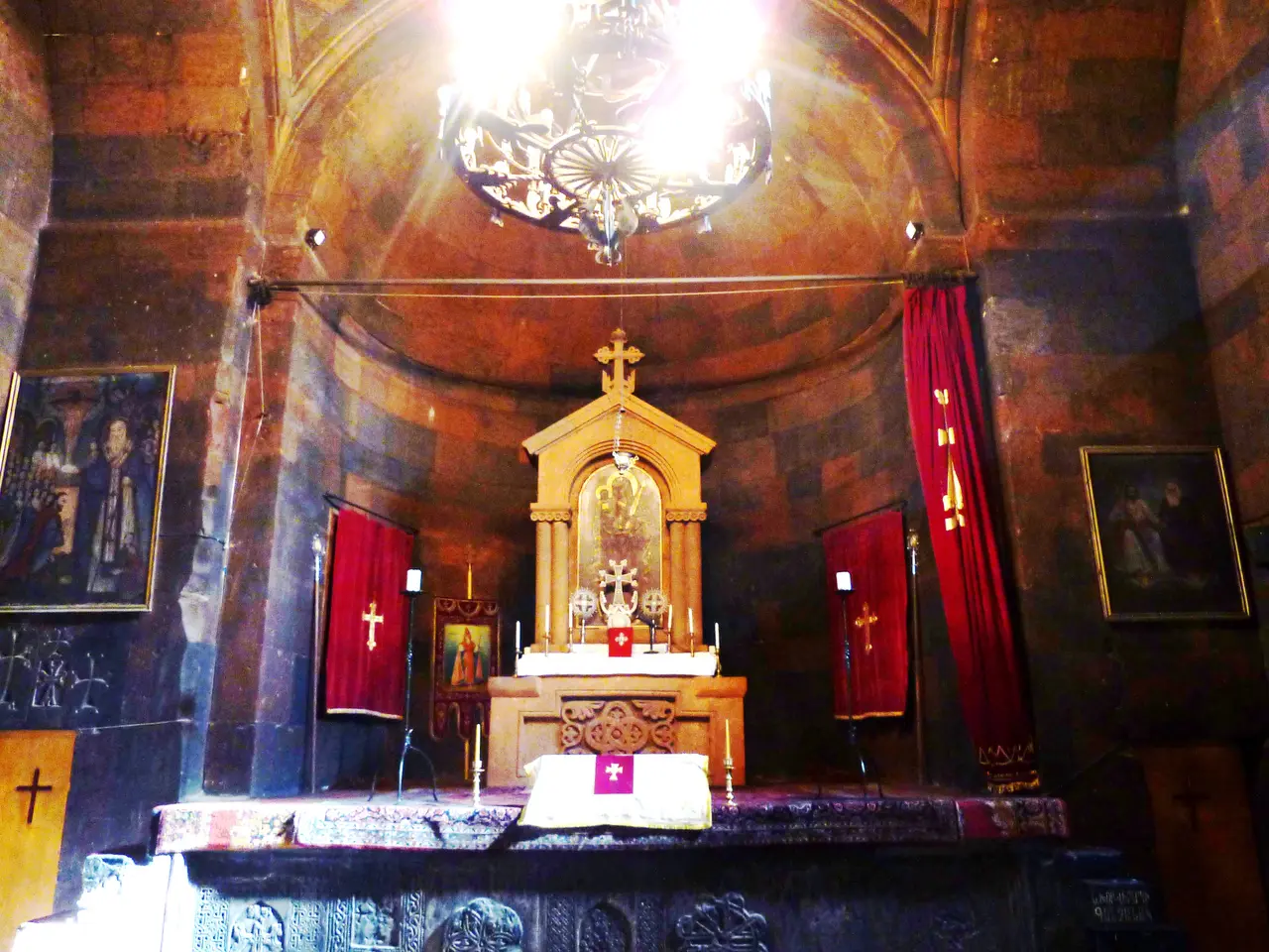 The dim interior of the Surb Astvatsatsin church at Khor Virap, with its stone altar, red hangings and carved khachkars lit by a hanging chandelier