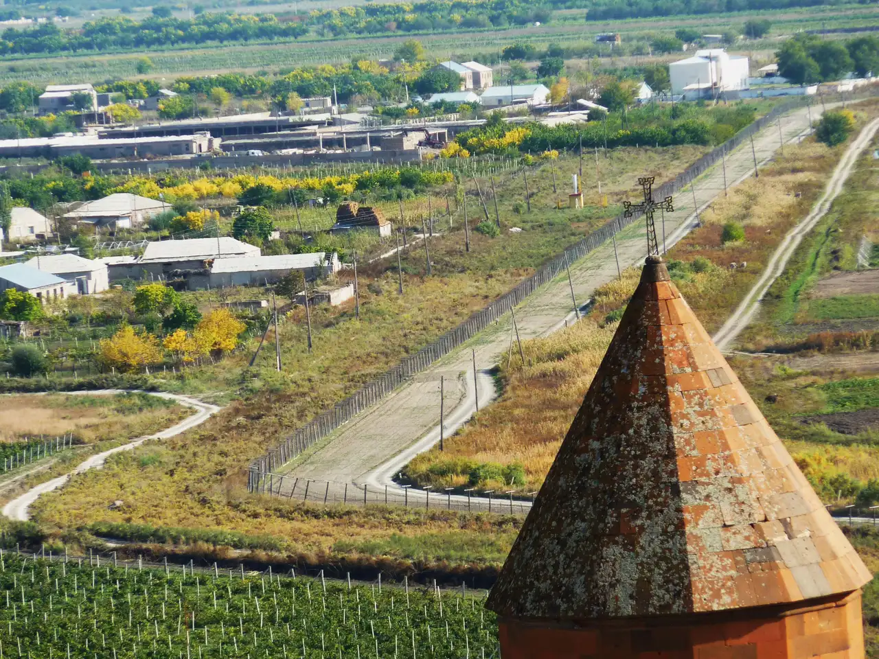 The cross-topped spire of Khor Virap above the fenced no-man's-land of the closed Armenia-Turkey border, with a village and vineyards beyond