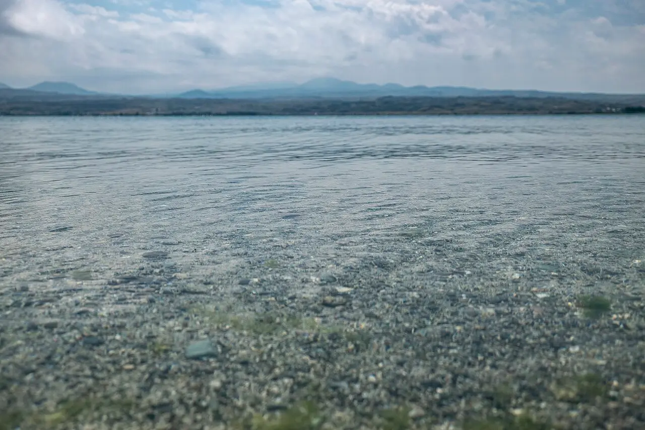 The clear, pebbly shallows of a Lake Sevan beach with the low mountain shore across the water under a cloudy sky