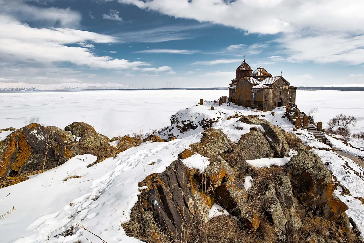 The stone Hayravank monastery on a snowy rocky headland above the frozen white surface of Lake Sevan under a blue sky with clouds