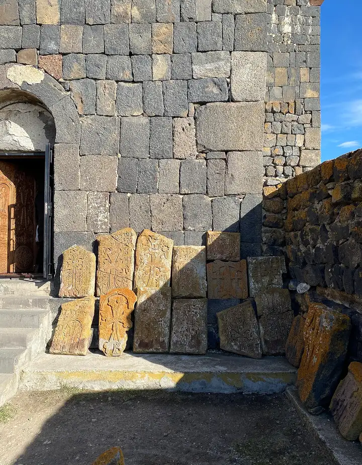 A row of carved khachkars (Armenian cross-stones) with orange lichen leaning against the basalt wall of a church at Sevanavank