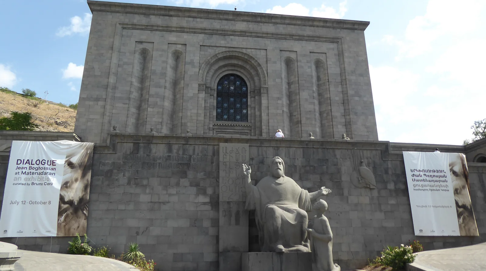 The grey stone facade of the Matenadaran in Yerevan seen from below, with the monumental sculpture of Mesrop Mashtots and his pupil at its base