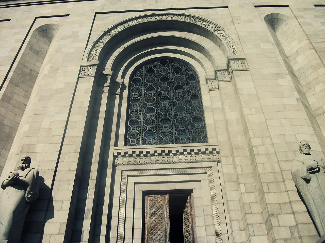 The arched stone entrance of the Matenadaran flanked by statues of Armenian scholars, with an ornate bronze door below