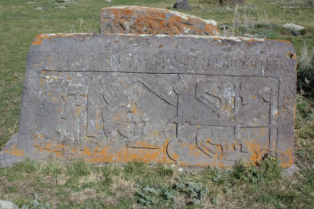 A flat lichen-covered tombstone at Noratus carved with figures and animals in a framed panel beneath a band of Armenian inscription