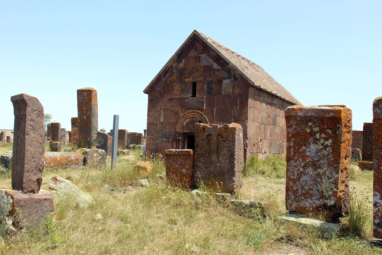A small red stone chapel with a pitched metal roof standing among lichen-covered khachkars in the summer grass at Noratus