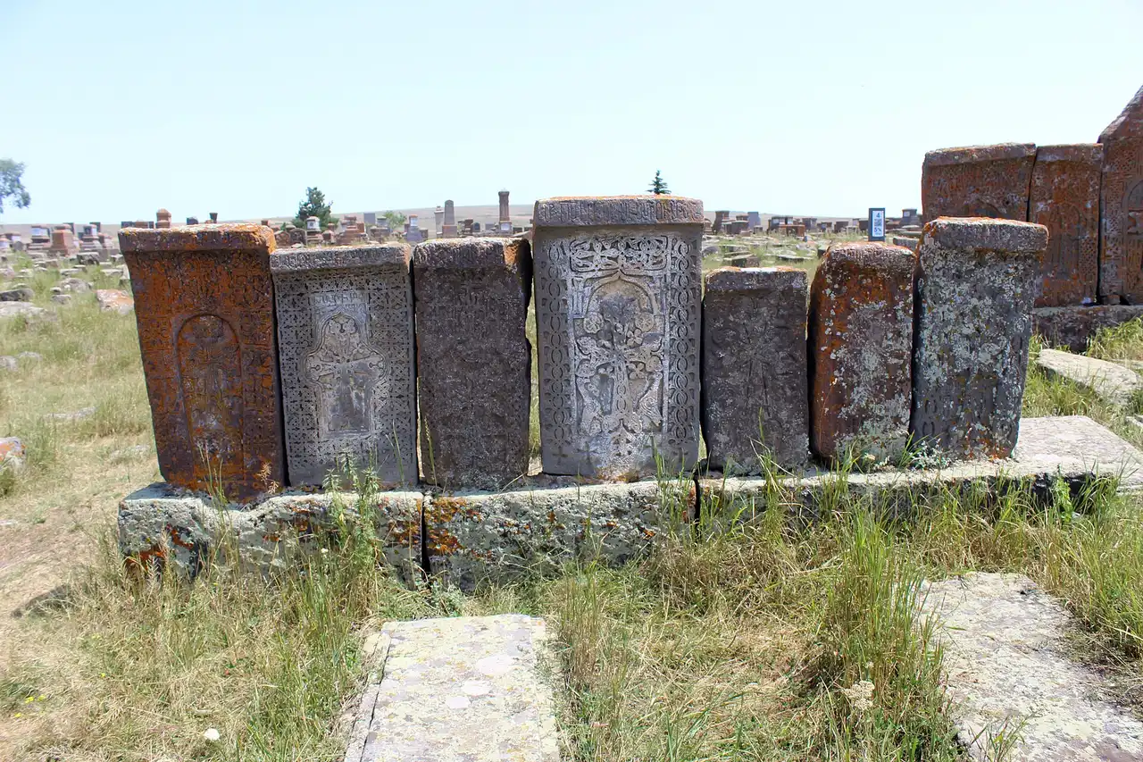 A row of medieval khachkars in orange lichen standing shoulder to shoulder on a stone plinth at Noratus, each carved with a different cross and interlace pattern