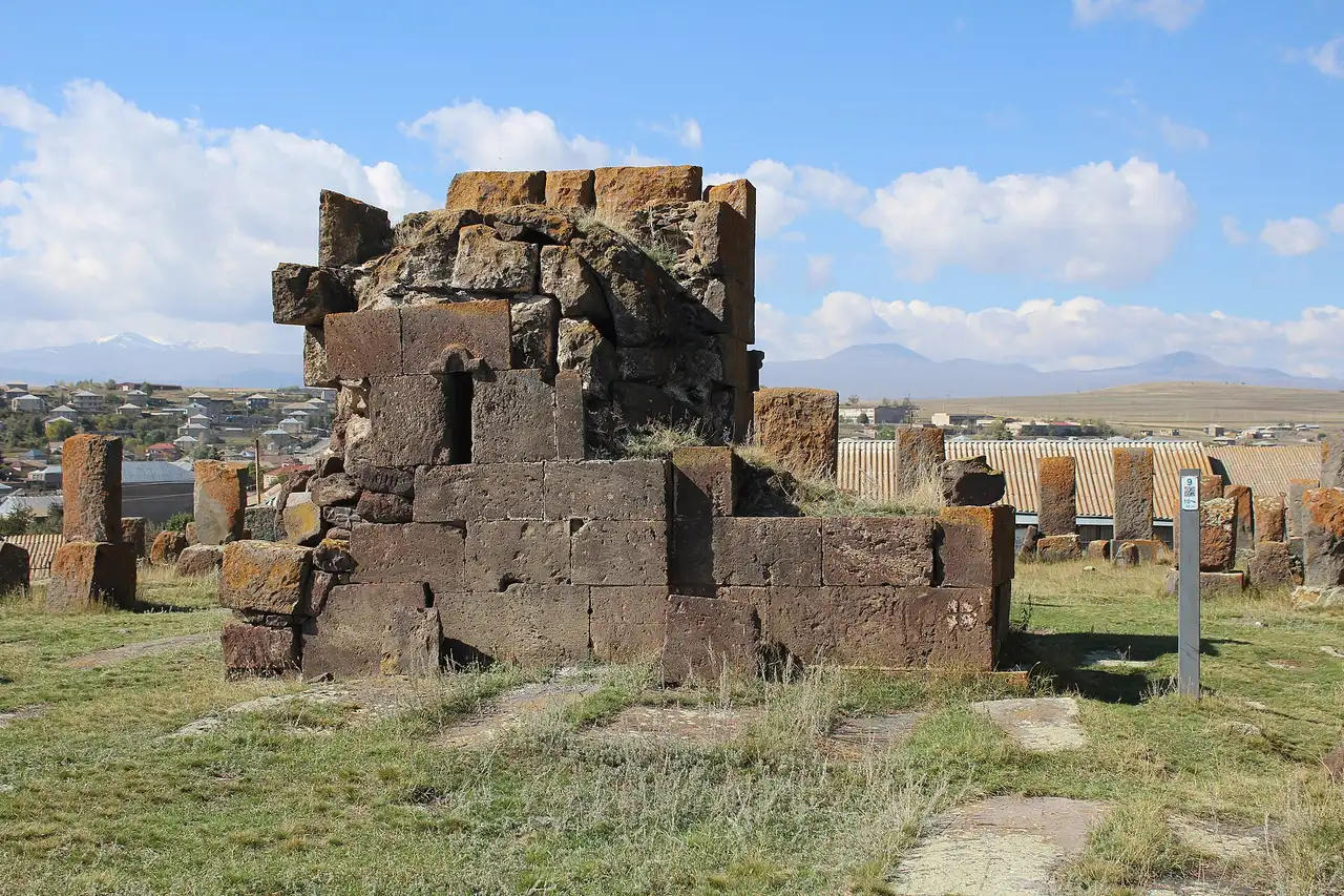 The stone ruin of a small building at the edge of Noratus cemetery, with khachkars around it and the village roofs and distant snow-streaked mountains behind