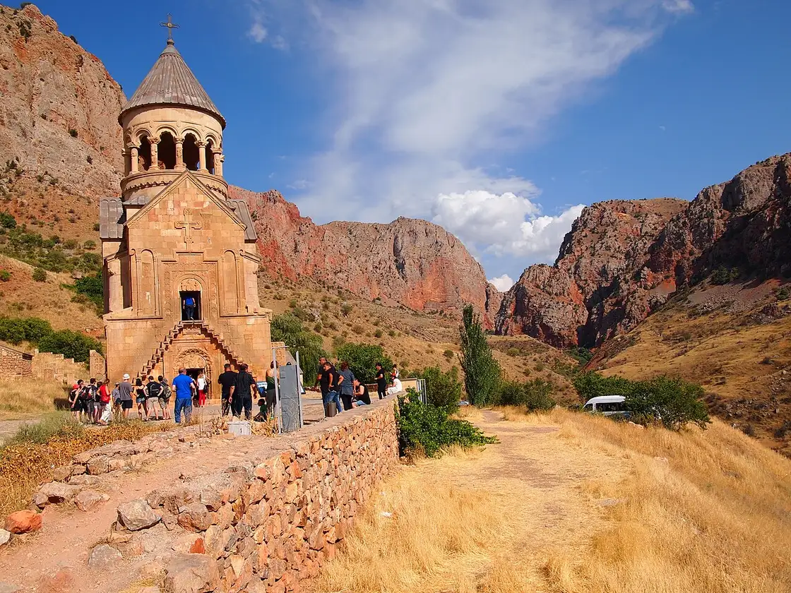 The two-storey Burtelashen church at Noravank with its lantern belfry, seen from the approach path with visitors gathered at its steps and the red canyon opening up behind
