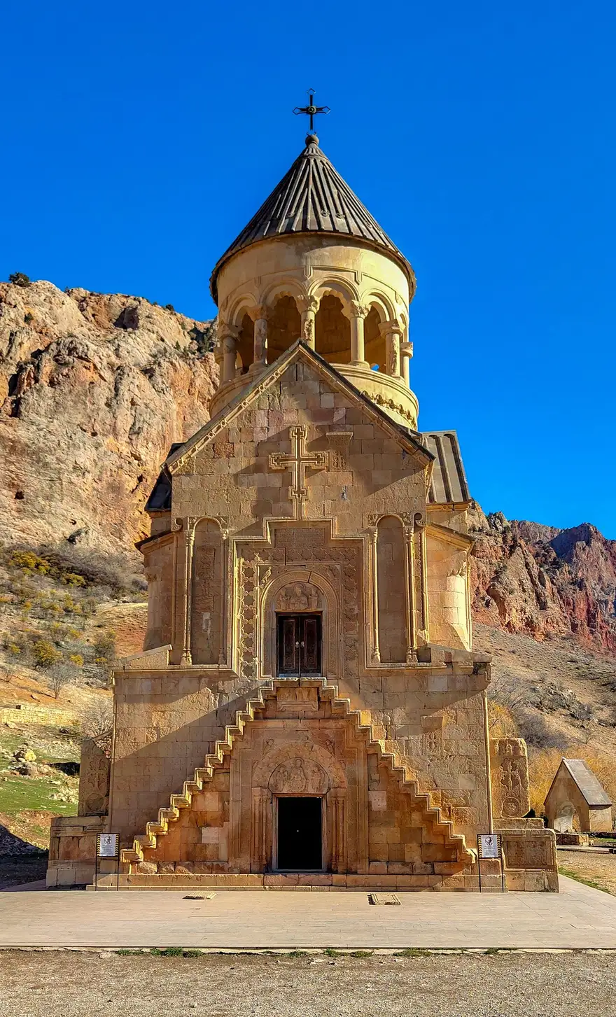 The west front of the two-storey Burtelashen church at Noravank, with two symmetrical flights of narrow stone steps projecting from the wall up to the first-floor doorway, red cliffs behind