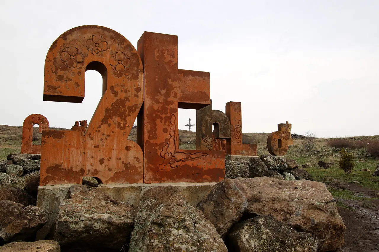 Two large rust-coloured tuff sculptures of Armenian letters carved with rosettes and interlace, standing on rough basalt boulders with a metal cross on the hill behind