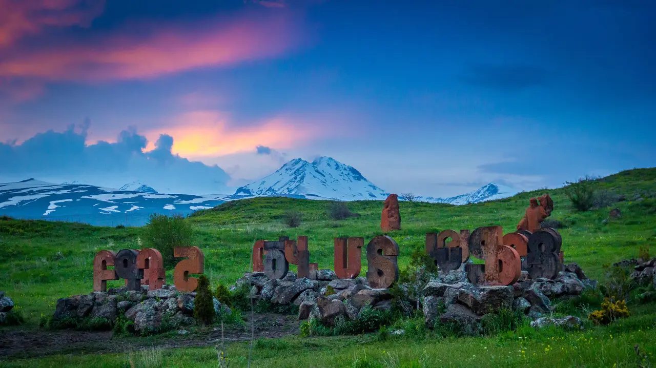 Giant carved stone letters of the Armenian alphabet standing in a green meadow at dusk with the snow-covered peaks of Mount Aragats behind