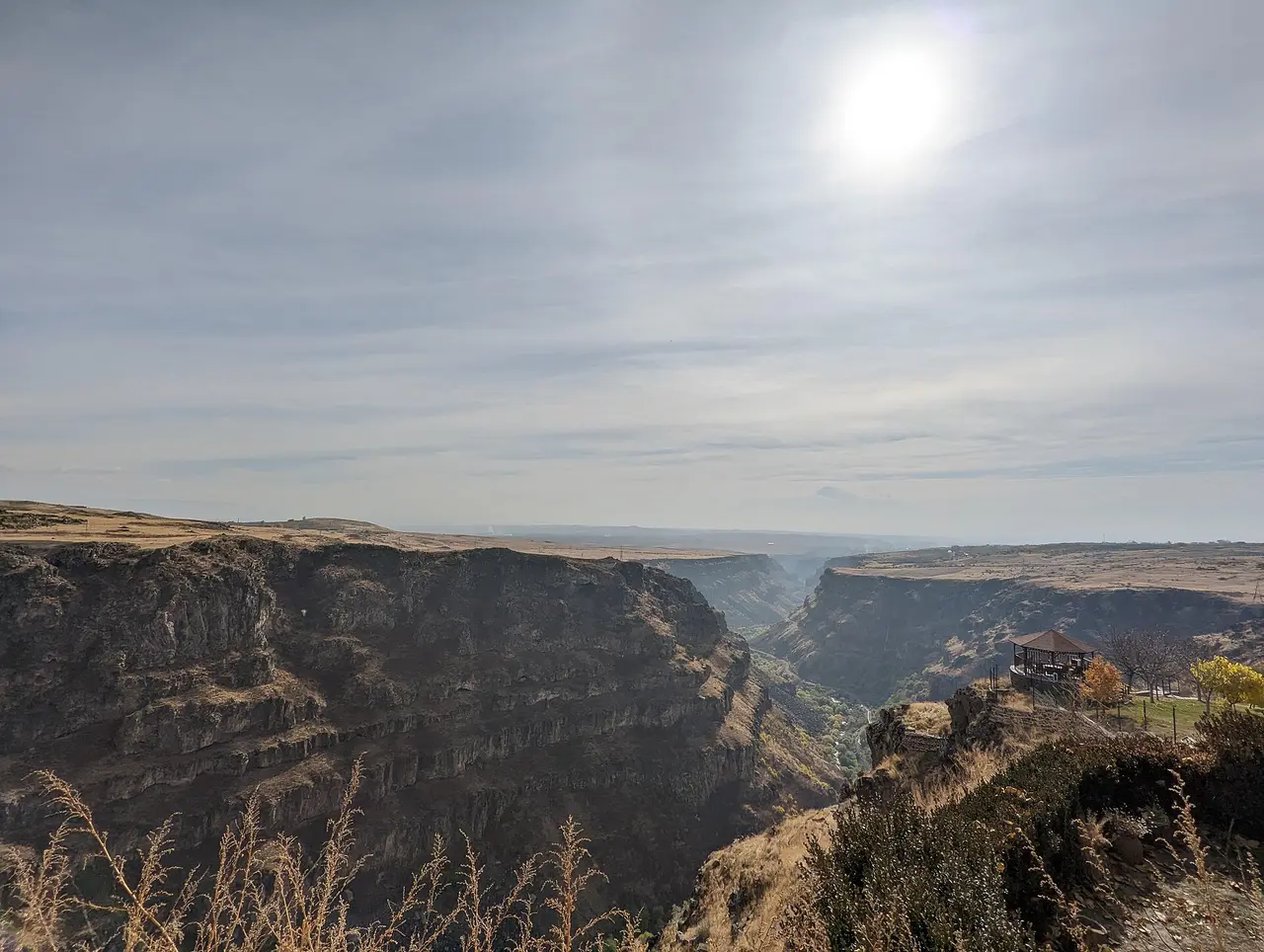 The stepped basalt cliffs of the Kasagh river gorge seen from the rim at Saghmosavan, with a covered viewpoint on a spur and the Ararat plain hazy in the distance