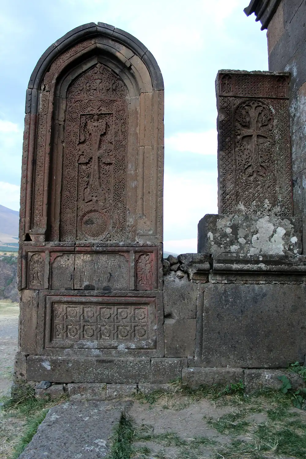 Two weathered khachkars, Armenian carved cross-stones, standing against the monastery wall at Saghmosavank with an inscription panel below and the gorge visible at the left