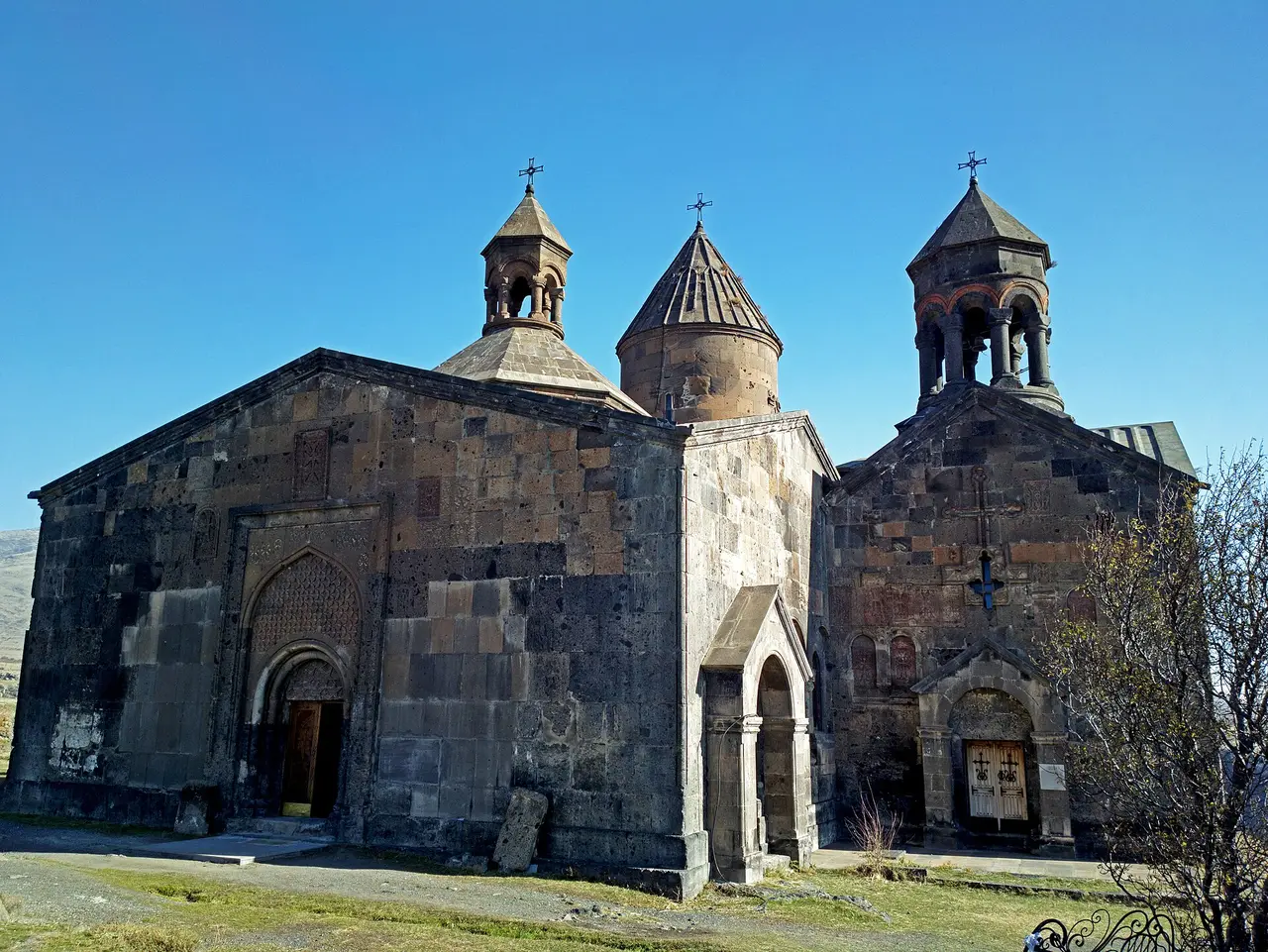 The dark tuff walls, carved portal and conical domes of the Saghmosavank monastic complex under a blue sky