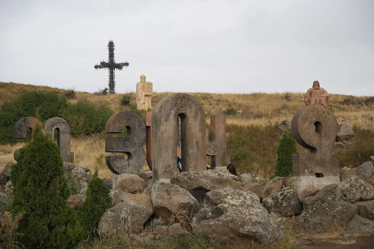 Stone Armenian letters in a dry summer meadow with seated statues of Armenian writers behind them and a tall openwork metal cross on the hillside