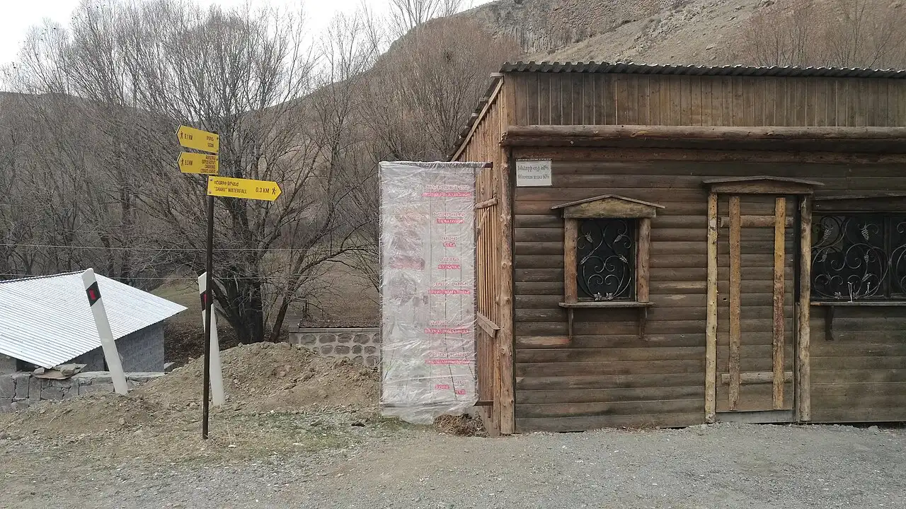 Yellow trail signposts and a wooden kiosk beside the gravel parking area at the Shaki waterfall on a bare winter afternoon