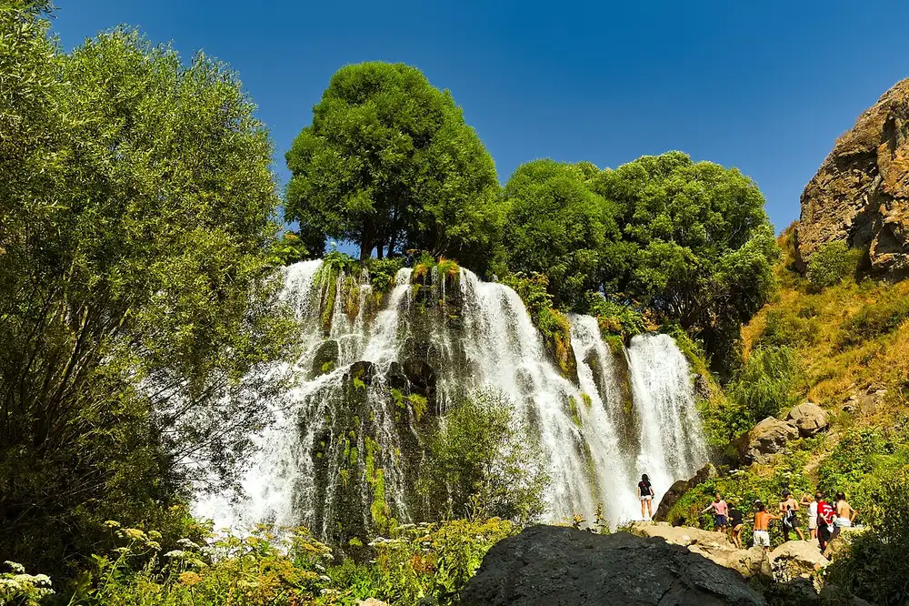 The Shaki waterfall in full summer flow with a group of visitors standing on the rocks at the foot of the falls