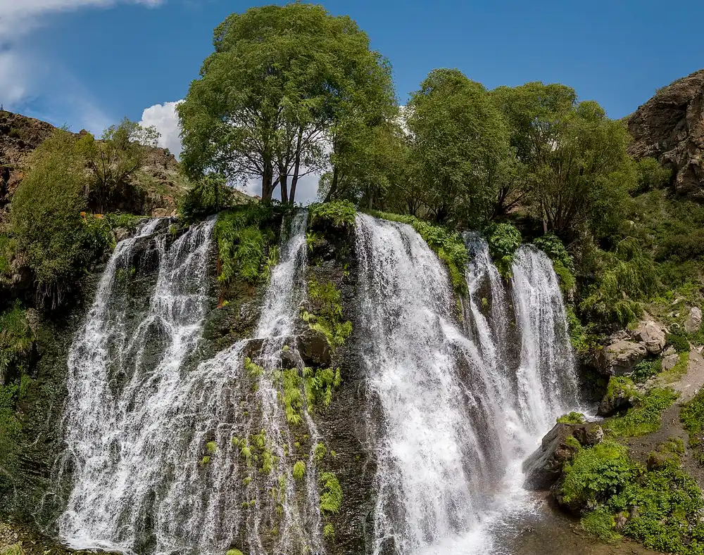 The whole Shaki waterfall seen from above the gorge in summer, water fanning across the full width of a green-topped basalt ledge