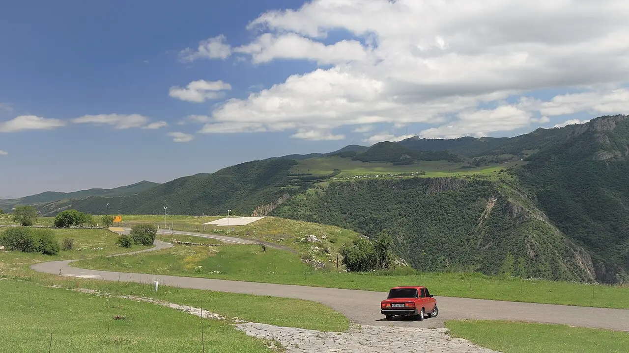 A red car on a quiet paved road above the green Vorotan gorge in southern Armenia, forested hills stretching to the horizon